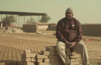 In this still from Pratik's documentary film, an Asian man sits atop a pile of bricks in a sandy, rather desolate landscape.