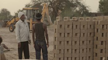 In this still from Pratik's documentary film, an older man in a turban speaks with a younger man whose back is to the camera. They stand by a large pile of bricks.