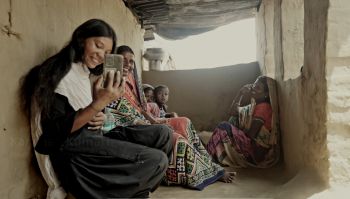 Asian women of different ages sit on the floor of an open concrete dwelling smiling and taking a selfie with a mobile phone