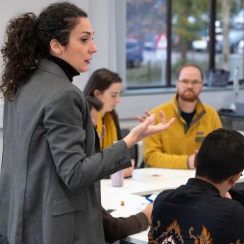 A woman facilitating a workshop, speaking to researchers sitting around tables.