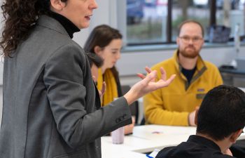 A woman facilitating a workshop, speaking to researchers sitting around tables.