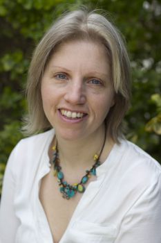 A portrait image of a smiling Perpetua Kirby wearing a white shirt and statement necklace in front of a blurred tree in the background