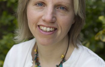 A portrait image of a smiling Perpetua Kirby wearing a white shirt and statement necklace in front of a blurred tree in the background