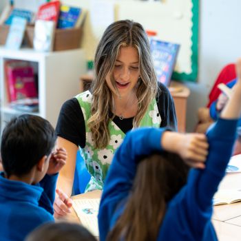 A trainee teacher sits with a group of primary school-aged pupils in a classroom setting