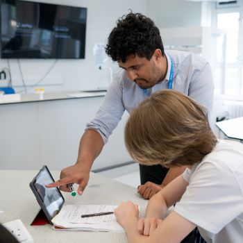 A young, male trainee teacher stands by a seated pupil and looks into his computer screen