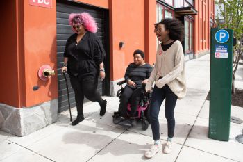 Three Black disabled people laugh together as they move along a windy pavement - one with a cane, one in a power wheelchair, and one walking beside them.