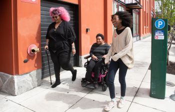 Three Black disabled people laugh together as they move along a windy pavement - one with a cane, one in a power wheelchair, and one walking beside them.