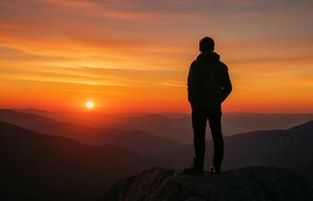 Person standing on a rocky cliff overlooking a vast mountain range at sunset, with vibrant orange and red hues filling the sky and the sun low on the horizon.