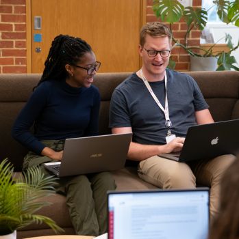 Two smiling researchers, a woman and a man, sit on a sofa, talking and sharing laptop screens.