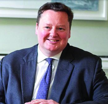 A professional portrait photograph of Richard Honey KC who is smiling whilst sat at a desk wearing a suit.