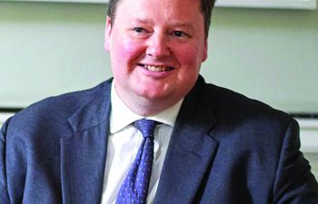 A professional portrait photograph of Richard Honey KC who is smiling whilst sat at a desk wearing a suit.