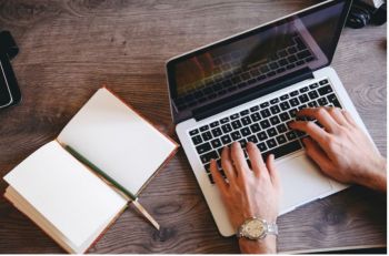 a photograph of a white person's hands typing on a silver laptop next to an open notebook and an pen, on a wooden table