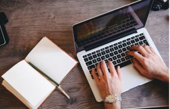 a photograph of a white person's hands typing on a silver laptop next to an open notebook and an pen, on a wooden table