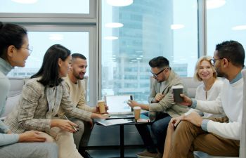 three men and three women sat in an office area talking and looking at a laptop.