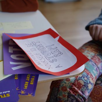 Posters on a table. One says 'Vote', another says ' Nominate yourself to be a Student Rep!'.  A person is sitting at the table but there is only their hand and legs in the frame..