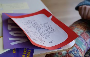 Posters on a table. One says 'Vote', another says ' Nominate yourself to be a Student Rep!'.  A person is sitting at the table but there is only their hand and legs in the frame..