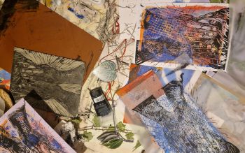 A messy craft table that has just been used with lino print supplies and illustrative lino prints of a person covering their face with their hands.