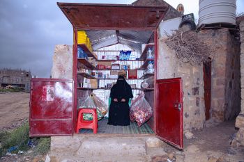 A small shop with open red doors, stocked with food and household items, with a woman standing inside.