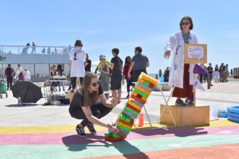 A science demonstration taking place on Brighton seafront. A speaker in a lab coat gestures as a volunteer topples a tower of blocks. In the background, a small crowd surrounds another speaker.