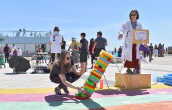 A science demonstration taking place on Brighton seafront. A speaker in a lab coat gestures as a volunteer topples a tower of blocks. In the background, a small crowd surrounds another speaker.