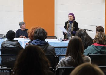 Janna Eliot in a purple headscarf reads from a paper at a microphone and an audience listens in a room with white walls and a bright orange stripe.