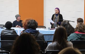 Janna Eliot in a purple headscarf reads from a paper at a microphone and an audience listens in a room with white walls and a bright orange stripe.