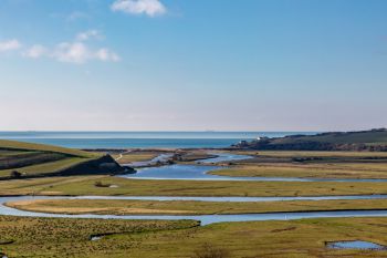 Meandering Cuckmere River flowing through green fields toward the sea under a clear blue sky.