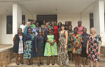 Around sixteen staff members stand together on steps outside a building at the University Of Ghana, they are smiling at the camera. The group is positioned in two rows in front of an entrance with pillars and open doors.