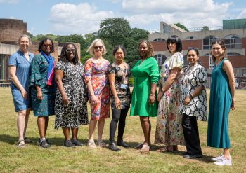 Eight staff members stand side by side on a grassy lawn outdoors, smiling at the camera. University Of Sussex buildings, and some trees are visible in the background under a blue sky.