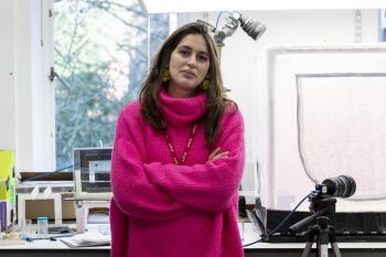 A member of staff stands in a laboratory with their arms crossed, looking at the camera. They are wearing a bright pink jumper and a staff lanyard. Laboratory equipment, including cameras and experimental apparatus, is visible on benches around them