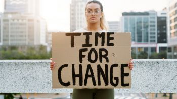 An image of a woman with glasses and a ponytail, standing in the centre of the frame and facing the camera, holding a cardboard placard that reads 