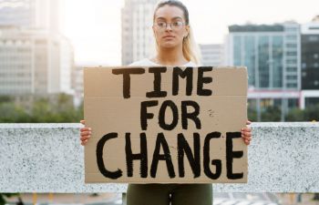 An image of a woman with glasses and a ponytail, standing in the centre of the frame and facing the camera, holding a cardboard placard that reads 