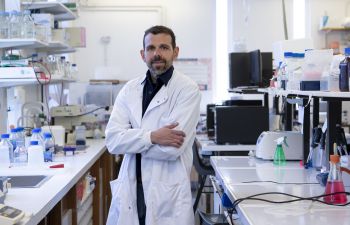 Professor Georgios Giamas wearing a white lab coat in a science laboratory in the University of Sussex. He is smiling and has his arms crossed.