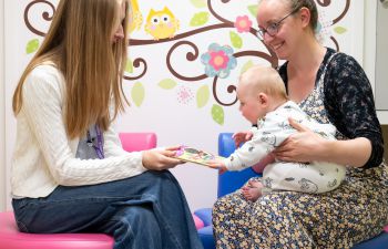 A baby sitting on a caregiver's lap reaches for a picture book