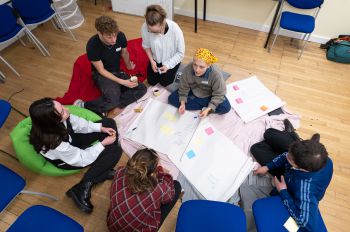 Students sitting on the floor, gathered around a piece of paper with pens.