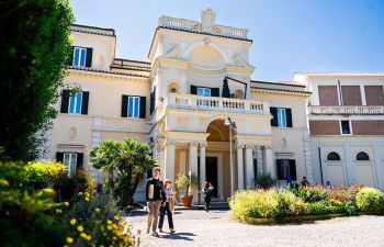 Two students walk in front of an impressive pale coloured building with columns and a balcony which is part of Luiss University in Rome, Italy.