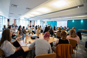 A large classroom-style workshop with many seated attendees using laptops, facing a panel of speakers at the front. Bright room with modern desks and windows.