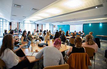 A large classroom-style workshop with many seated attendees using laptops, facing a panel of speakers at the front. Bright room with modern desks and windows.