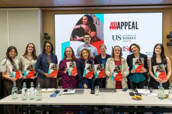 A photograph of 9 smiling women all holding copies of the new guide with one smiling man standing behind them. In the background is a large slide showing logos of the funders and workers behind the guide.