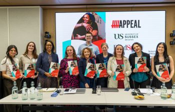 A photograph of 9 smiling women all holding copies of the new guide with one smiling man standing behind them. In the background is a large slide showing logos of the funders and workers behind the guide.