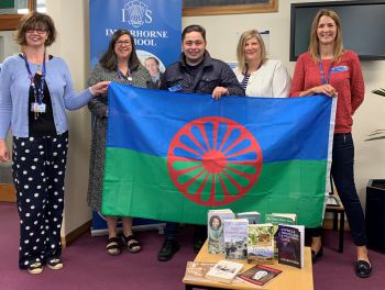 Five people standing indoors holding a GRT flag, with books and resources displayed in front of them to mark Gypsy, Roma and Traveller History Month.