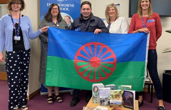 Five people standing indoors holding a GRT flag, with books and resources displayed in front of them to mark Gypsy, Roma and Traveller History Month.