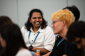 Photograph of a student with long hair, a watch and a collared shirt smiling at the camera, with other students sat blurred out in the foreground around them.
