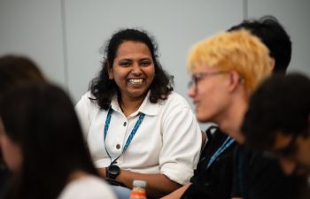 Photograph of a student with long hair, a watch and a collared shirt smiling at the camera, with other students sat blurred out in the foreground around them.