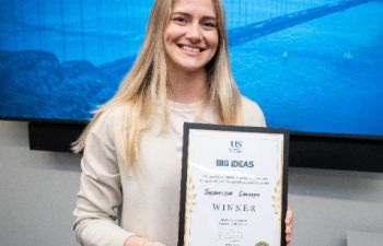 Image of Jacqueline holding up a certificate in front of a 'Big Ideas Contest' blue screen