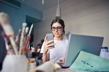 Image of a woman with dark hair and glasses looking at a phone, with a laptop and paintbrushes in the foreground.