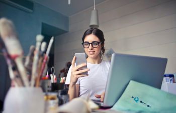 Image of a woman with dark hair and glasses looking at a phone, with a laptop and paintbrushes in the foreground.