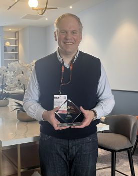 A standing Dr Roesch is shown smiling at the camera and holding his award from BISA in an informal setting which could be his living room.