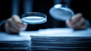 out of focus hands holding two magnifying glasses above a pile of papers