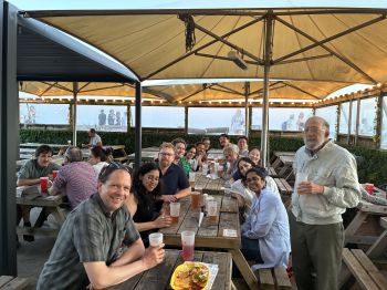 A large group of conference participants sit around wooden picnic tables under a covered outdoor seating area, smiling toward the camera with drinks on the table.